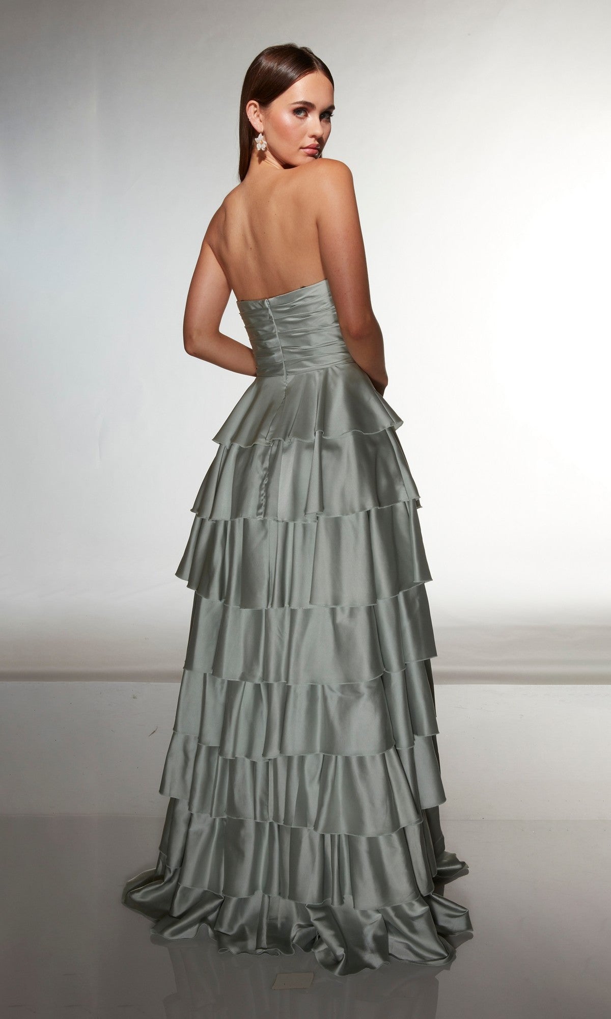 A woman stands with her back to the camera in the Alyce 61987 long prom dress, a strapless, floor-length silver satin gown featuring tiered ruffles, against a plain background.